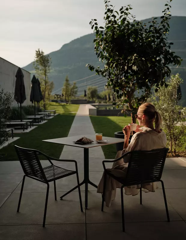 Frau mit Getränk auf der Terrasse mit Blick über Liegewiese und Berge